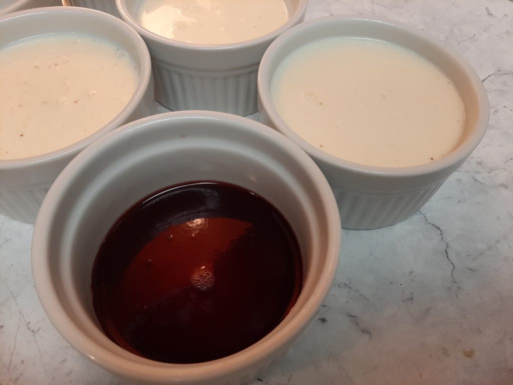 Port Wine gelee in the bottom of a ceramic ramekin surrounded by ramekins with cardamom panna cotta on a marble counter