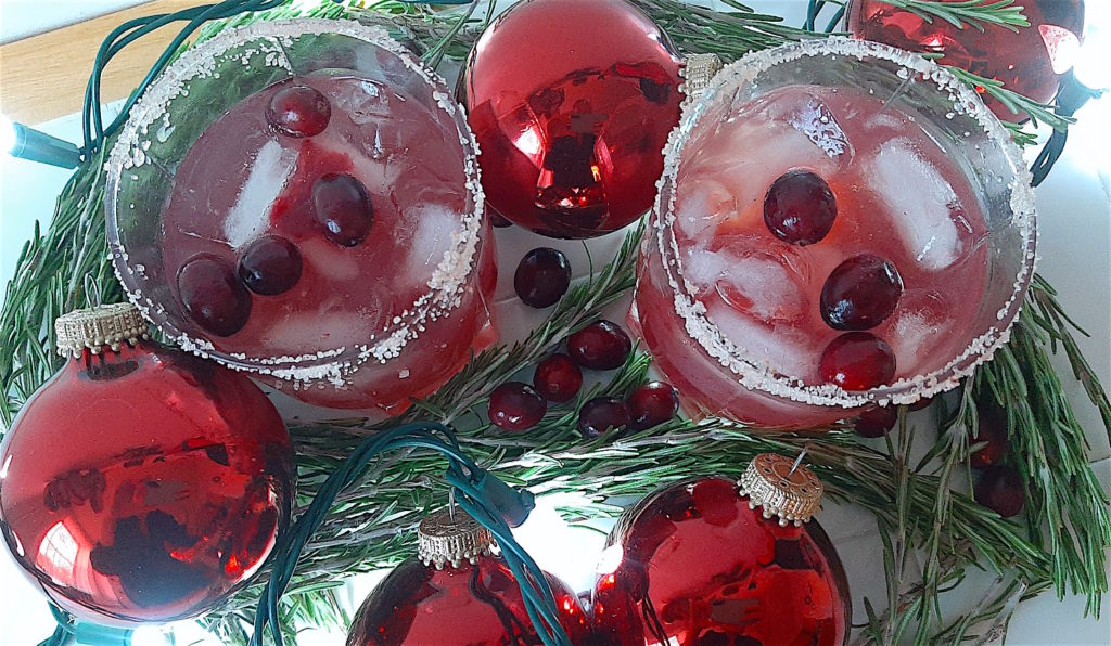 Cranberry Margaritas on the rocks with tequila, triple sec, cranberry juice, lime juice, and cranberries surrounded by rosemary, cranberries, and Christmas ornaments and lights. Christmas cocktail.