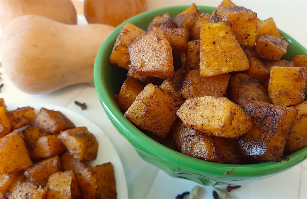 Roasted butternut squash in green bowl and plate with cardamom and winter spices surrounded with butternut squash, cloves, and cardamom seeds.