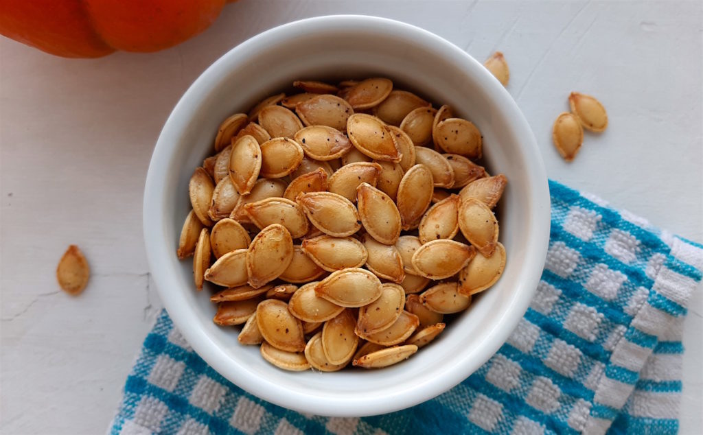 Roast Spaghetti squash seeds in a white ramekin on a blue checked cloth with a pumpkin the background. They are easy to make, just like any other roast winter squash seeds.