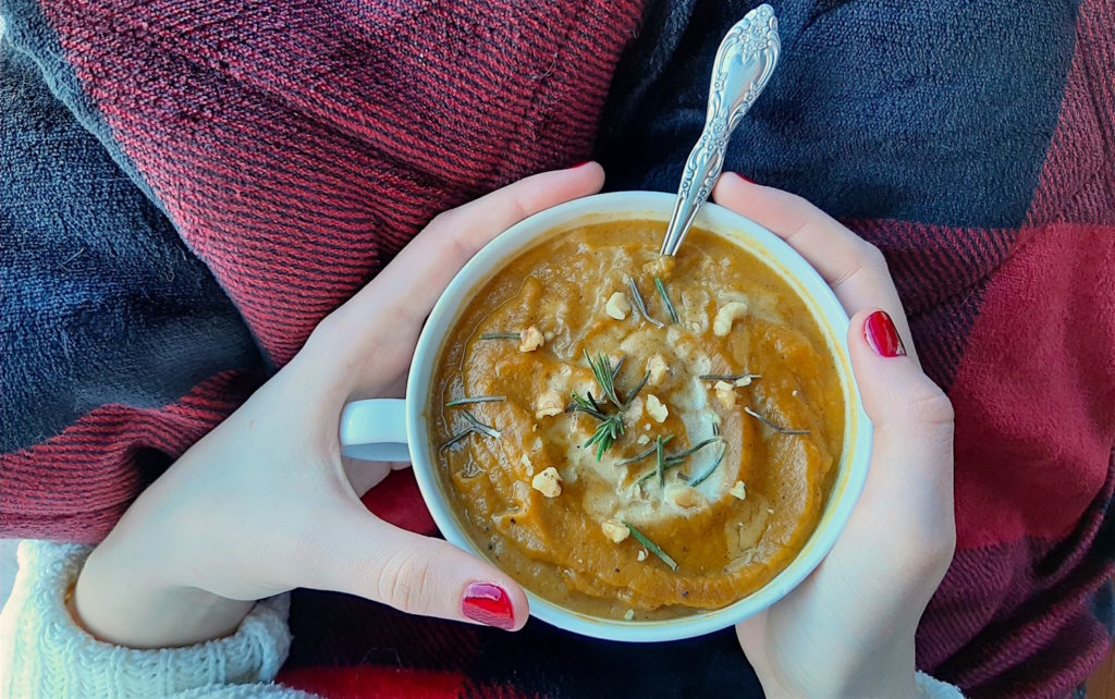 Woman with red nails holding a cup of brown butter butternut squash soup with cream, walnuts, and rosemary in a window seat with a red plaid blanket and white sweater. A cozy Sunday.