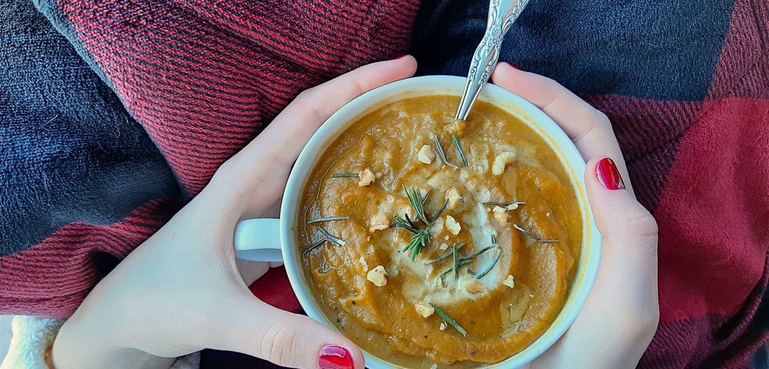 Butternut squash soup with brown butter, topped with rosemary, cream, and pumpkin seeds in a cup with a silver spoon held by a woman's hands with red nail polish over a red and black plaid blanket