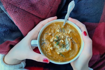 Butternut squash soup with brown butter, topped with rosemary, cream, and pumpkin seeds in a cup with a silver spoon held by a woman's hands with red nail polish over a red and black plaid blanket