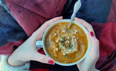 Butternut squash soup with brown butter, topped with rosemary, cream, and pumpkin seeds in a cup with a silver spoon held by a woman's hands with red nail polish over a red and black plaid blanket