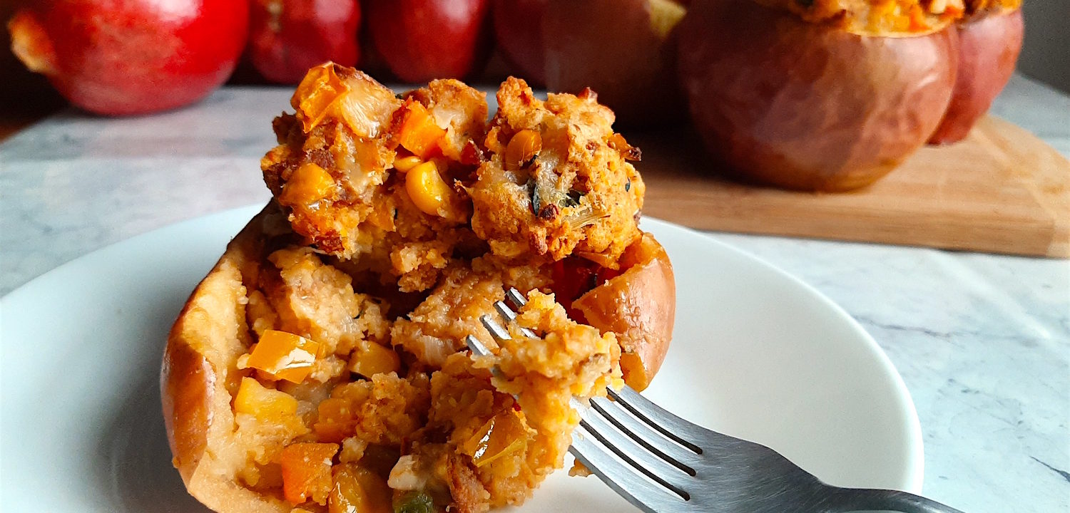 Sliced savory stuffed apple with Chorizo and Cornbread stuffing on a plate with a fork, more stuffed and whole apples in the background. Sugar with Spice Blog