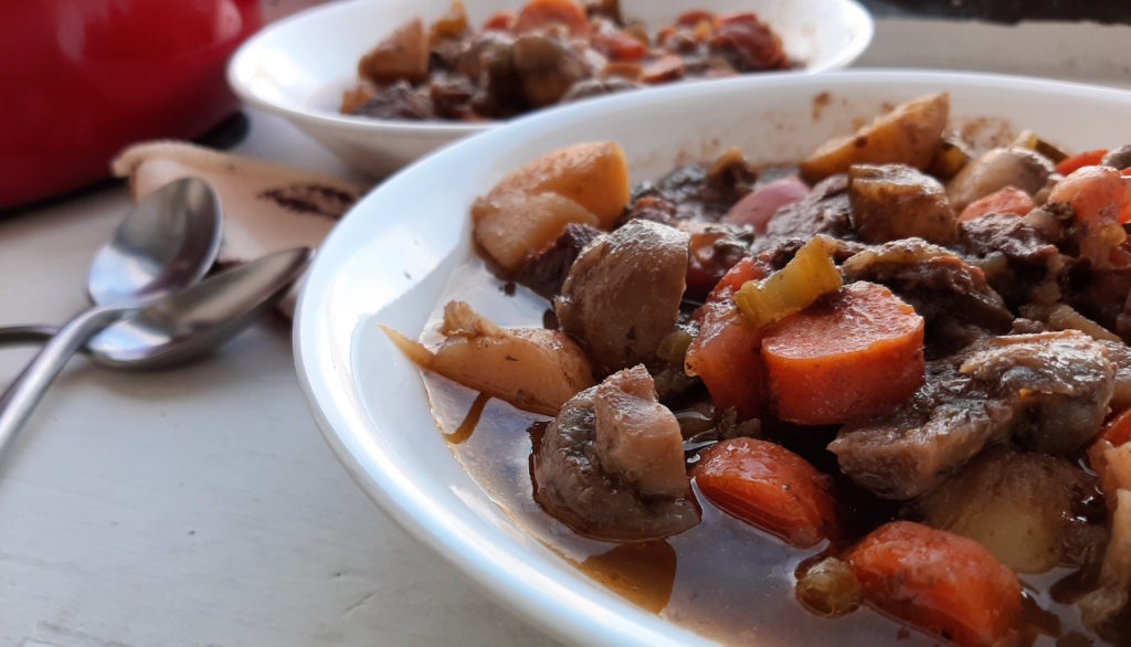 Beef stew with carrots, mushrooms, potatoes, and tomatoes in two white bowls with silver spoons next to a big red pot