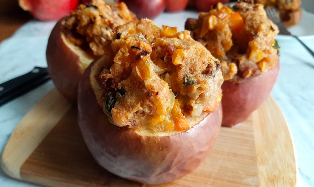 Three savory stuffed apples with Chorizo and Cornbread stuffing on a wooden cutting board with apples and a sliced apple in the background. Sugar with Spice Blog