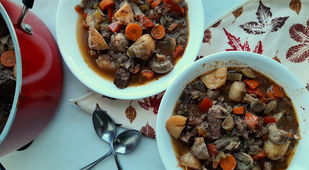 Beef Stew with carrots, mushrooms, and potatoes in two white bowls with silver spoons on top of a leaf towel. Next to a red nonstick pot with stew