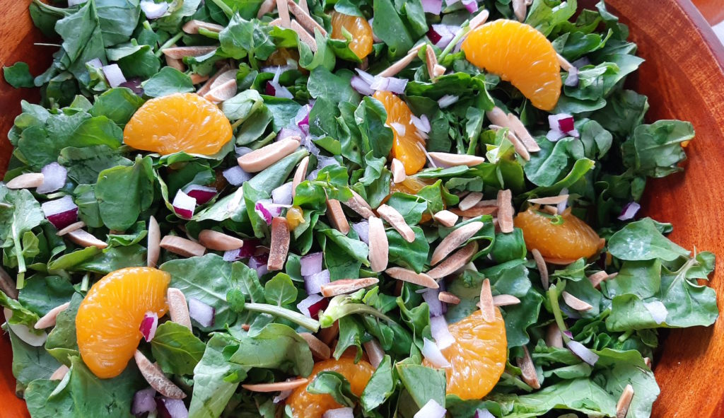 Watercress with mandarin oranges, toasted almonds, red onions in a dark wood bowl, tossed together