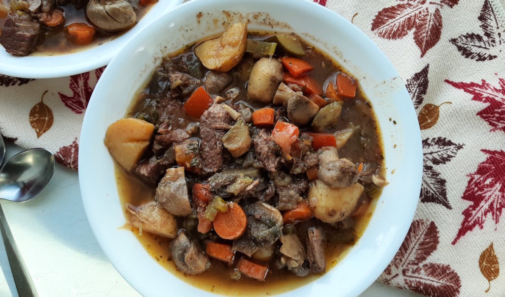Beef stew with carrots, tomatoes, potatoes, mushrooms, celery in two white bowls with silver spoons over a leaf patterned kitchen towel