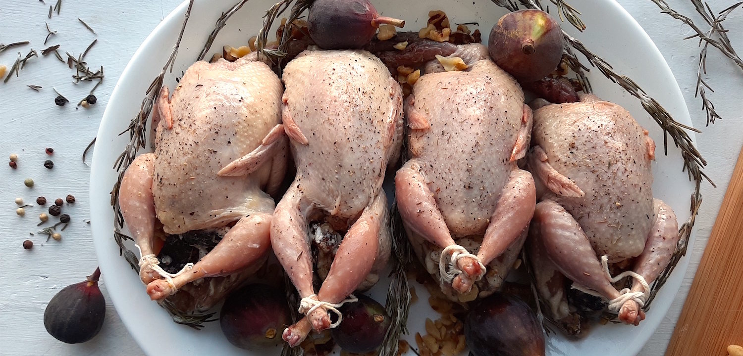 4 raw quail stuffed with figs, walnuts, and goat chese sprinkled with salt, olive oil, thyme, and pepper. Quail on a white plate surrounded by rosemary, figs, peppercorns and walnuts.