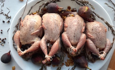 4 raw quail stuffed with figs, walnuts, and goat chese sprinkled with salt, olive oil, thyme, and pepper. Quail on a white plate surrounded by rosemary, figs, peppercorns and walnuts.