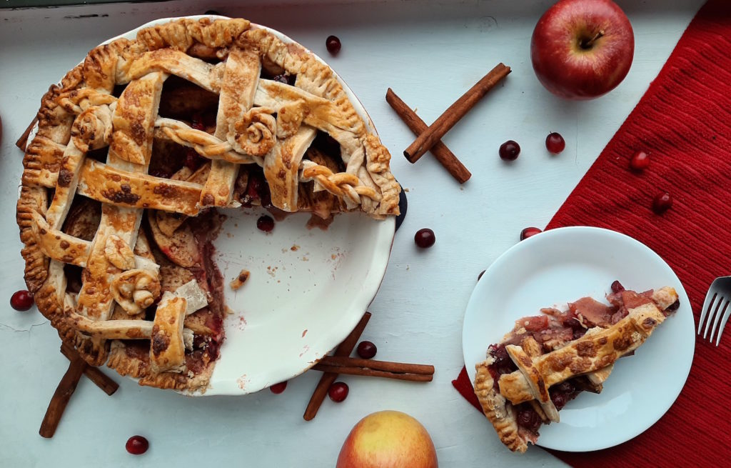 Cooked spiced cranberry apple pie with spiced all-butter pie crust, sliced on a plate and fork with apples, cinnamon, cranberries, and red towel. Sugar with Spice Blog.