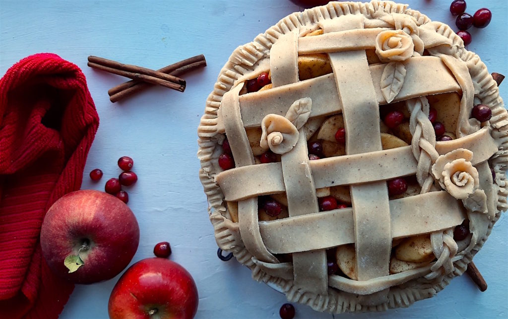 Spiced Cranberry Apple Pie with spiced all-butter pie crust raw in a ceramic dish with braided lattice crust and rose designs, surrounded by apples, cranberries, cinnamon sticks, and a red tea towel. Sugar with Spice Blog