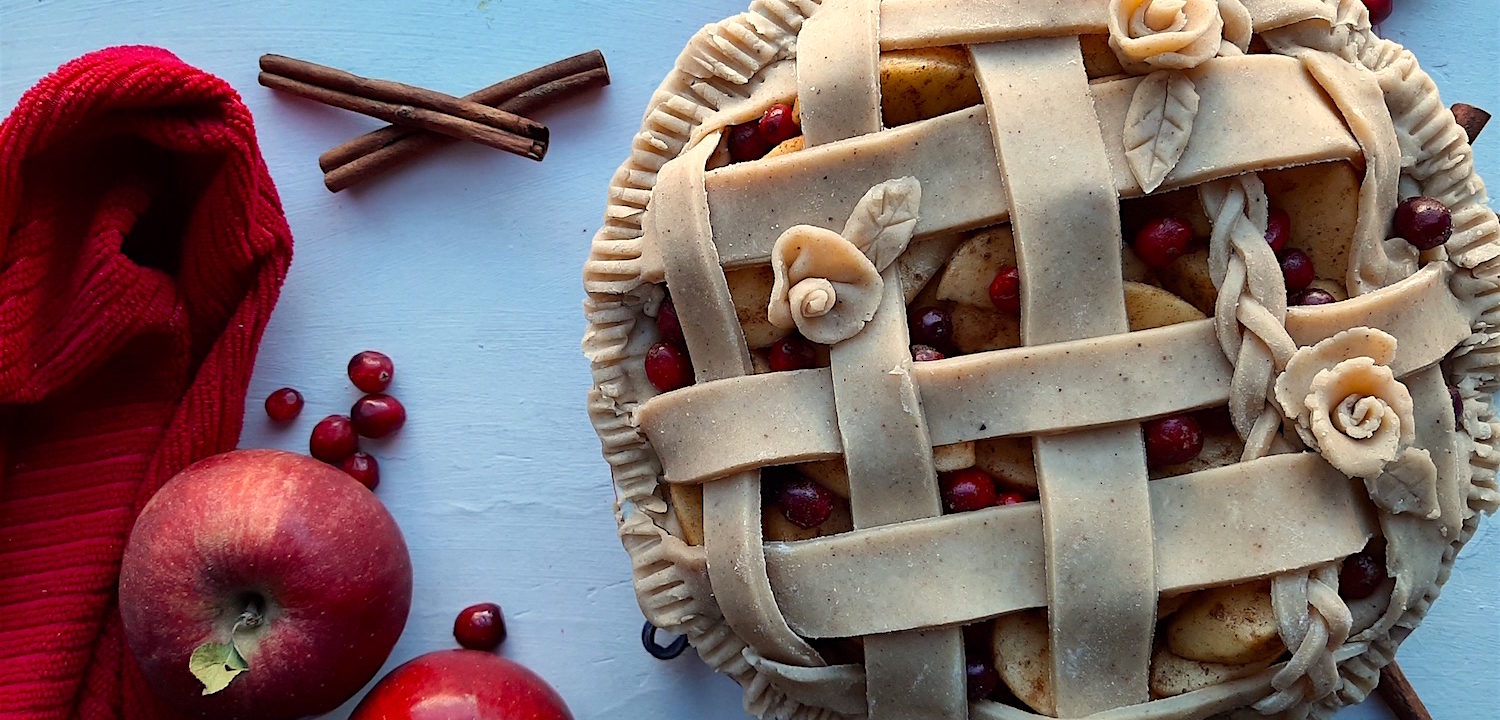 Spiced Cranberry Apple Pie with spiced all-butter pie crust raw in a ceramic dish with braided lattice crust and rose designs, surrounded by apples, cranberries, cinnamon sticks, and a red tea towel. Sugar with Spice Blog