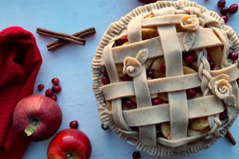 Spiced Cranberry Apple Pie with spiced all-butter pie crust raw in a ceramic dish with braided lattice crust and rose designs, surrounded by apples, cranberries, cinnamon sticks, and a red tea towel. Sugar with Spice Blog