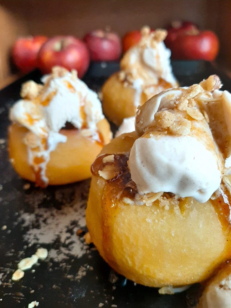 Sherry Poached Apples with Crumb Topping filling and Cinnamon Gelato covered in a sherry glaze on a baking dish with apples in the background. Sugar with Spice Blog.