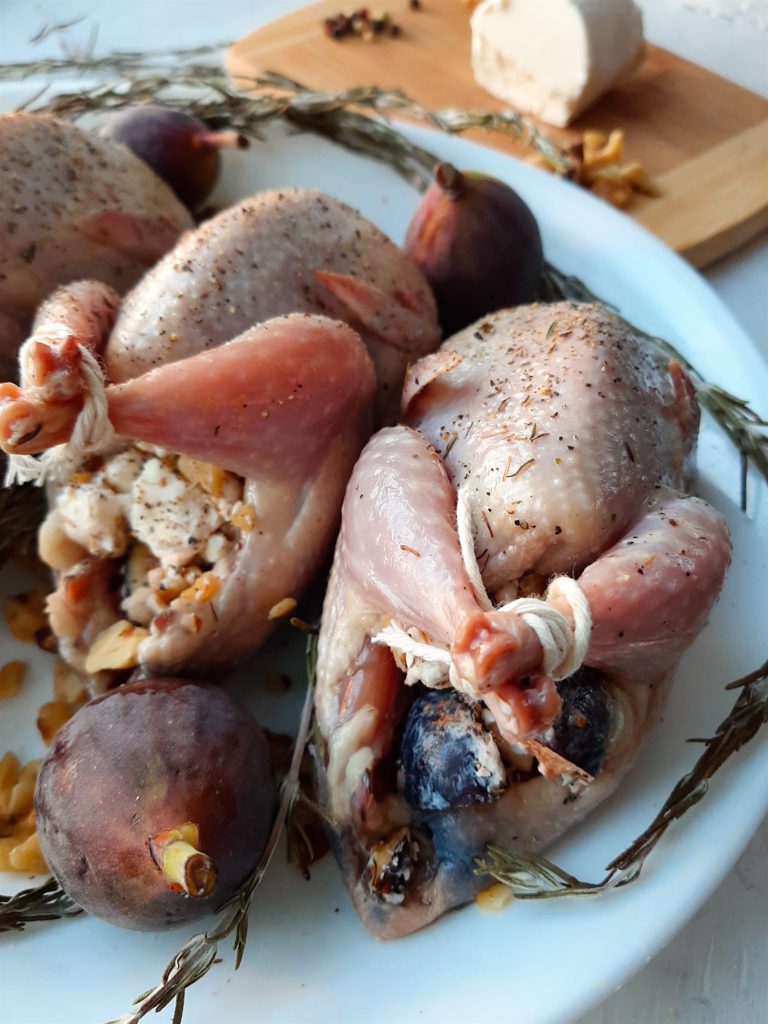 Roast stuffed quail stuffed with figs, walnuts, and goat chese sprinkled with salt, olive oil, thyme, and pepper. Quail on a white plate surrounded by rosemary, figs, peppercorns and walnuts with wooden cutting board with goat cheese in the background.