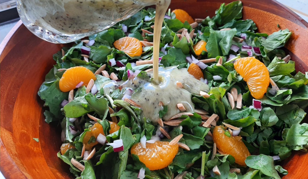 Watercress with mandarin oranges, toasted almonds, red onions in a dark wood bowl with a glass cup pouring lemon poppy seed dressing over it.