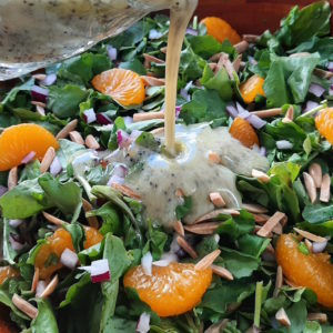 Watercress with mandarin oranges, toasted almonds, red onions in a dark wood bowl with a glass cup pouring lemon poppy seed dressing over it.
