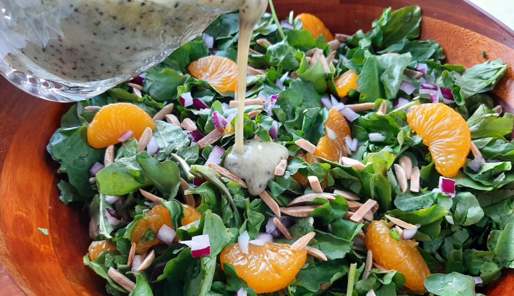 Watercress with mandarin oranges, toasted almonds, red onions in a dark wood bowl with a glass cup pouring lemon poppy seed dressing over it.