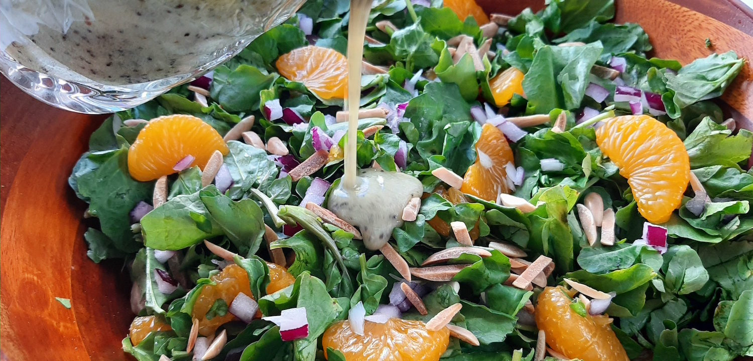 Watercress and Mandarin Orange Salad with Toasted Almonds, Red onions, and Lemon Poppy Seed Dressing in a wooden bowl. Lemon Poppy Seed dressing being poured from a glass over salad.