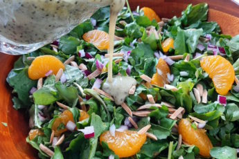 Watercress and Mandarin Orange Salad with Toasted Almonds, Red onions, and Lemon Poppy Seed Dressing in a wooden bowl. Lemon Poppy Seed dressing being poured from a glass over salad.