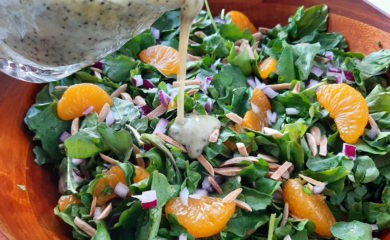 Watercress and Mandarin Orange Salad with Toasted Almonds, Red onions, and Lemon Poppy Seed Dressing in a wooden bowl. Lemon Poppy Seed dressing being poured from a glass over salad.