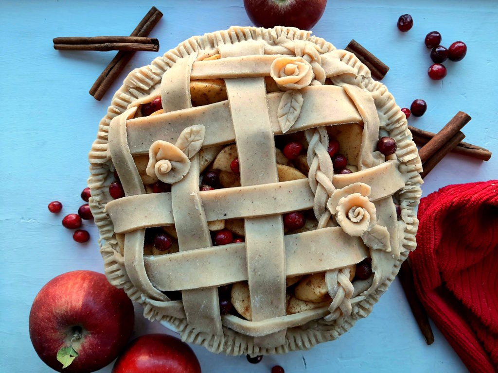 Spiced Cranberry Apple Pie with spiced all-butter pie crust raw in a ceramic dish with braided lattice crust and rose designs, centered, surrounded by apples, cranberries, cinnamon sticks, and a red tea towel. Sugar with Spice Blog