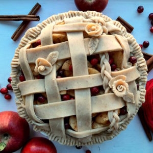 Spiced Cranberry Apple Pie with spiced all-butter pie crust raw in a ceramic dish with braided lattice crust and rose designs, centered, surrounded by apples, cranberries, cinnamon sticks, and a red tea towel. Sugar with Spice Blog