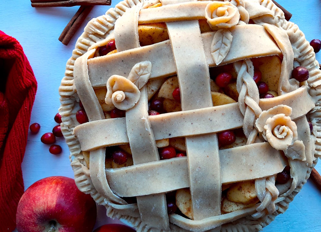 Spiced Cranberry Apple Pie with spiced all-butter pie crust raw in a ceramic dish with braided lattice crust and rose designs, close up, surrounded by apples, cranberries, cinnamon sticks, and a red tea towel. Sugar with Spice Blog
