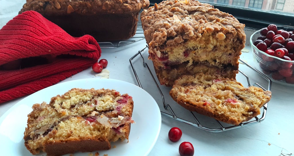 Two Cranberry Orange coffee cake loaves cooling on a window sill with a bowl of fresh cranberries and a red tea towel. One loaf is sliced. Sugar with Spice