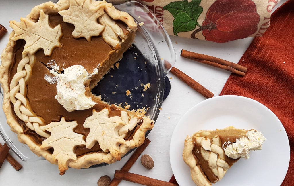 Fresh pumpkin pie made with fresh pumpkin and all-butter pie crust in a glass dish on a blue oven mitt, sliced on a plate surrounded by orange tea towels, pumpkin tea kitchen towel, cinnamon sticks and nutmeg. Sugar with Spice Blog