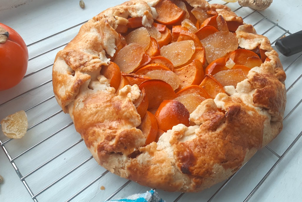 Persimmon and ginger galette on a cooling rack surrounded by persimmons, ginger, and cardamom seeds with a blue checked dish towel. Sugar with Spice Blog.