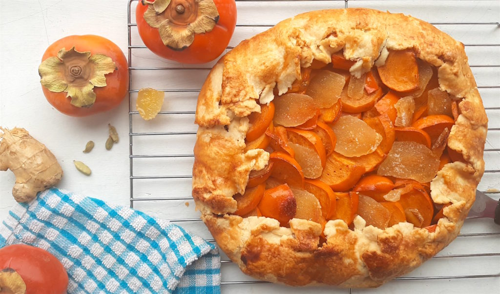 Persimmon and ginger galette on a cooling rack surrounded by persimmons, ginger, and cardamom seeds with a blue checked dish towel. Sugar with Spice Blog.