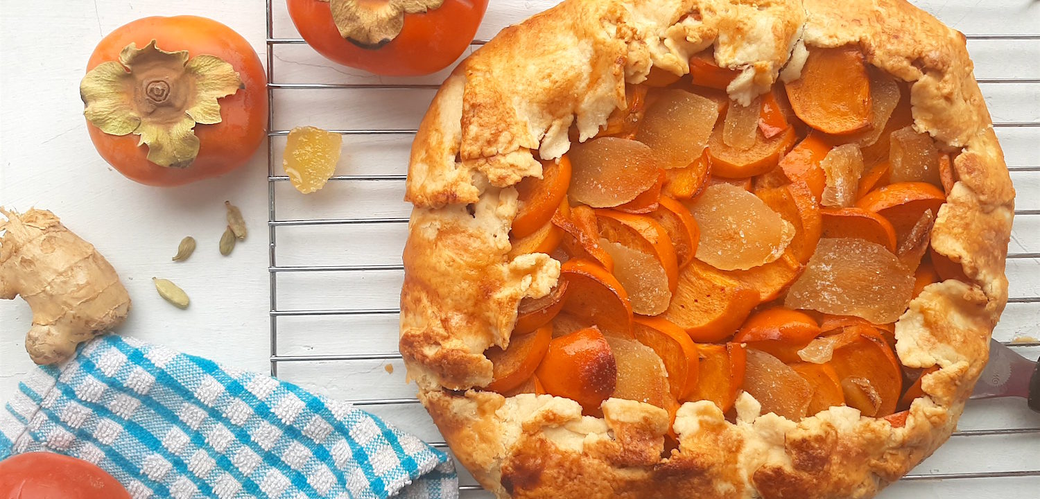 Persimmon and ginger galette on a cooling rack surrounded by persimmons, ginger, and cardamom seeds with a blue checked dish towel. Sugar with Spice Blog.