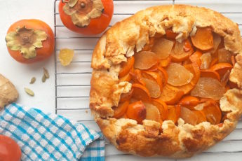 Persimmon and ginger galette on a cooling rack surrounded by persimmons, ginger, and cardamom seeds with a blue checked dish towel. Sugar with Spice Blog.