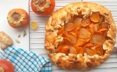 Persimmon and ginger galette on a cooling rack surrounded by persimmons, ginger, and cardamom seeds with a blue checked dish towel. Sugar with Spice Blog.