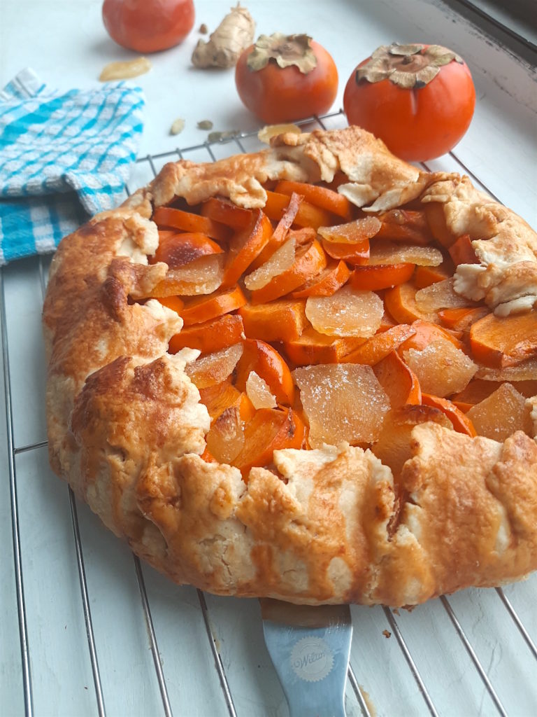 Persimmon and ginger galette on a cooling rack surrounded by persimmons, ginger, and cardamom seeds with a blue checked dish towel. Sugar with Spice Blog.