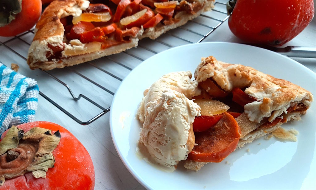 Two persimmon and ginger galettes, one sliced in half, on a cooling rack surrounded by persimmons, ginger, and cardamom seeds with a blue checked dish towel. One slice of persimmon ginger galette on a white dish with honeycomb ice cream. Sugar with Spice Blog.
