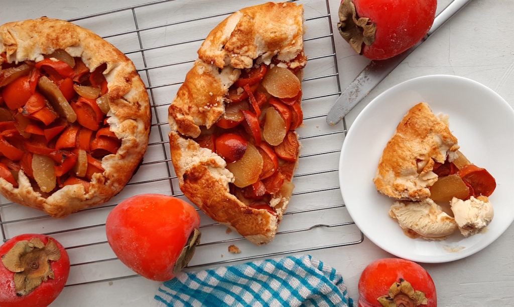 Two persimmon and ginger galettes, one sliced in half, on a cooling rack surrounded by persimmons, ginger, and cardamom seeds with a blue checked dish towel. One slice of persimmon ginger galette on a white dish with honeycomb ice cream. Sugar with Spice Blog.