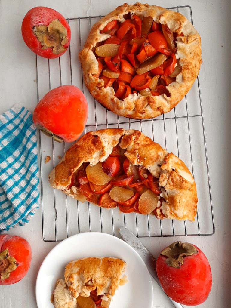 Two persimmon and ginger galettes, one sliced in half, on a cooling rack surrounded by persimmons, ginger, and cardamom seeds with a blue checked dish towel. One slice of persimmon ginger galette on a white dish with honeycomb ice cream. Sugar with Spice Blog.