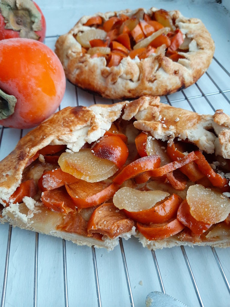 Sliced Persimmon and ginger galette on a cooling rack with a full galette in the background surrounded by hachiya persimmons. Sugar with Spice Blog.
