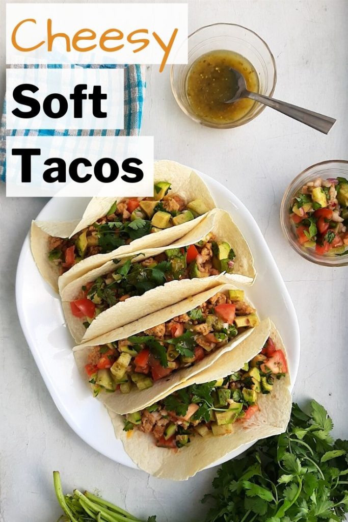Pinterest image for cheesy homemade soft tacos with flour tortillas. Four soft tacos with ground chicken, refried beans, cheddar cheese, shallots, tomatoes, cucumber, avocado, jalapeños, cilantro. Salsa verde and more fresh relish to the side in pyrex bowls.