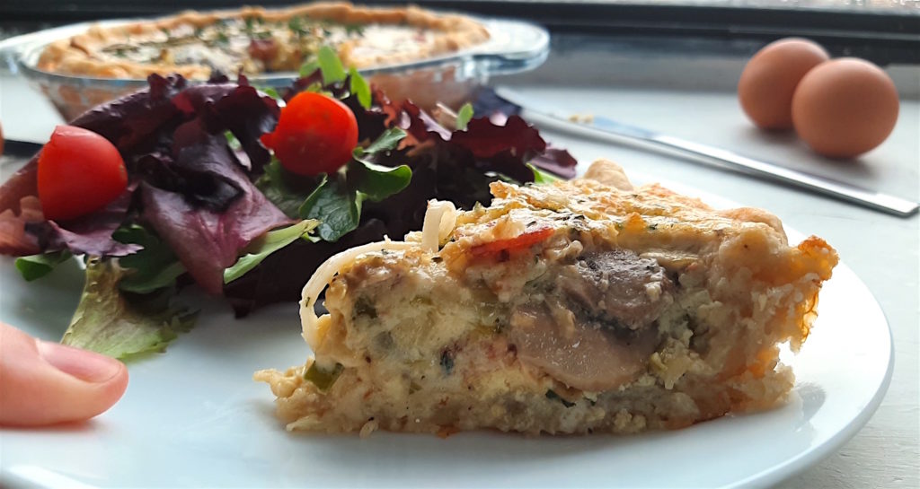 Mushroom, Leek, and Bacon Quiche slice on a white plate with salad being help by a woman's hand. Full quiche in a glass pan pan in the background. Surrounded by eggs and thyme stems
