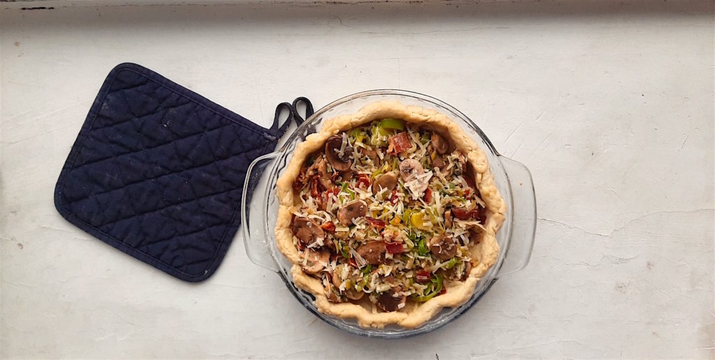 Mushroom, leek and bacon quiche with blind baked crust filled with sauteed mushrooms, leeks, and bacon in a glass pie dish and blue oven mit on a white background