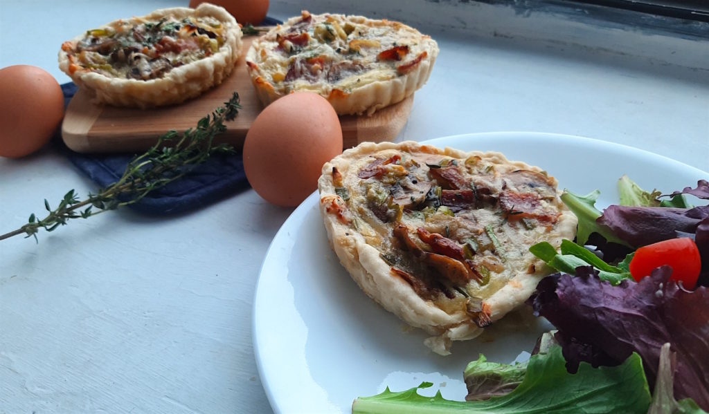 Mini heart-shaped mushroom, leek, and bacon quiche. One on a white plate with salad in the foreground. Two on a wooden cutting board and blue oven mit in the background. Eggs and thyme stems resting around.