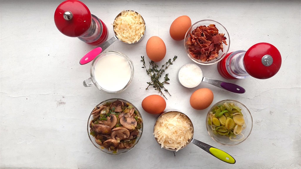 Ingredients for a mushroom, leek and bacon quiche. Salt and pepper shakers, milk, cream, sauteed mushrooms and leeks, 4 whole eggs, thyme stems, guyere, and parmesan on a white background