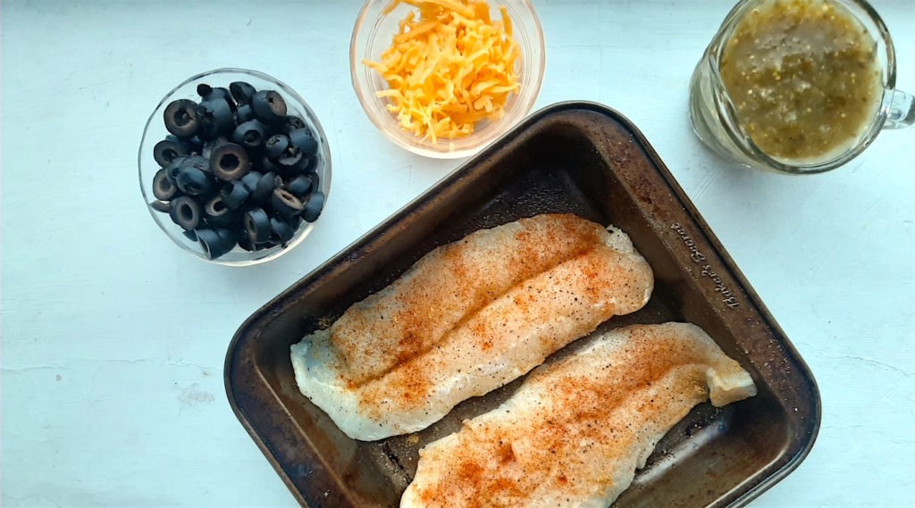 Basa filets seasoned with salt, pepper, and paprika in a casserole dish surrounded with bowls of black olives, cheddar cheese, and tomatillo sauce