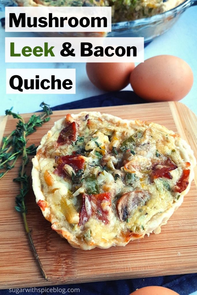 Heart-shaped mushroom, leek and bacon quiche on a wooden cutting board and blue oven mitt surrounded by thyme stems and whole eggs. Full quiche in glass pie dish in the background. Mushroom, leek and bacon quiche pinterest image. Sugar with Spice Blog.